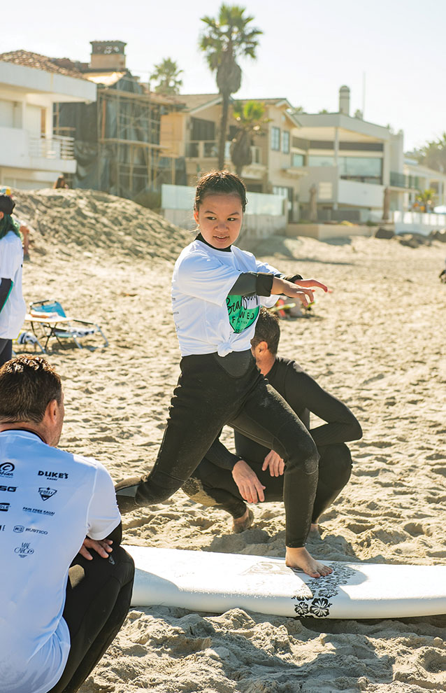 girl in white t-shirt and black neoprene suit practices standing up on surfboard on the sand