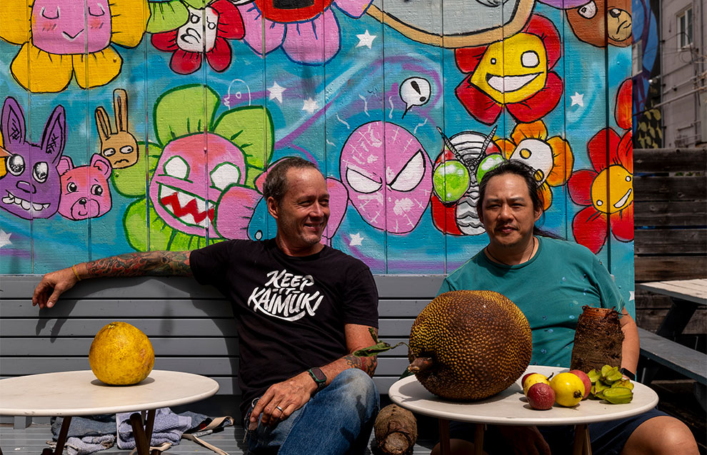two people sitting in front of a coloful mural, sitting beside a variety of fruits