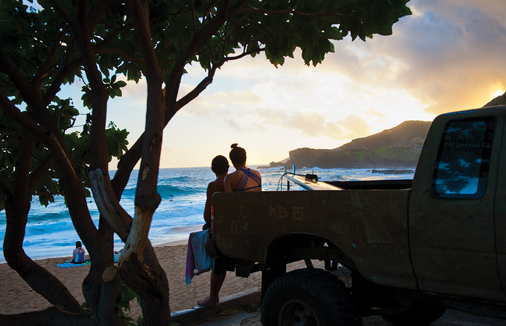 two people sitting at the back of a truck looking over to the sunset at the beach