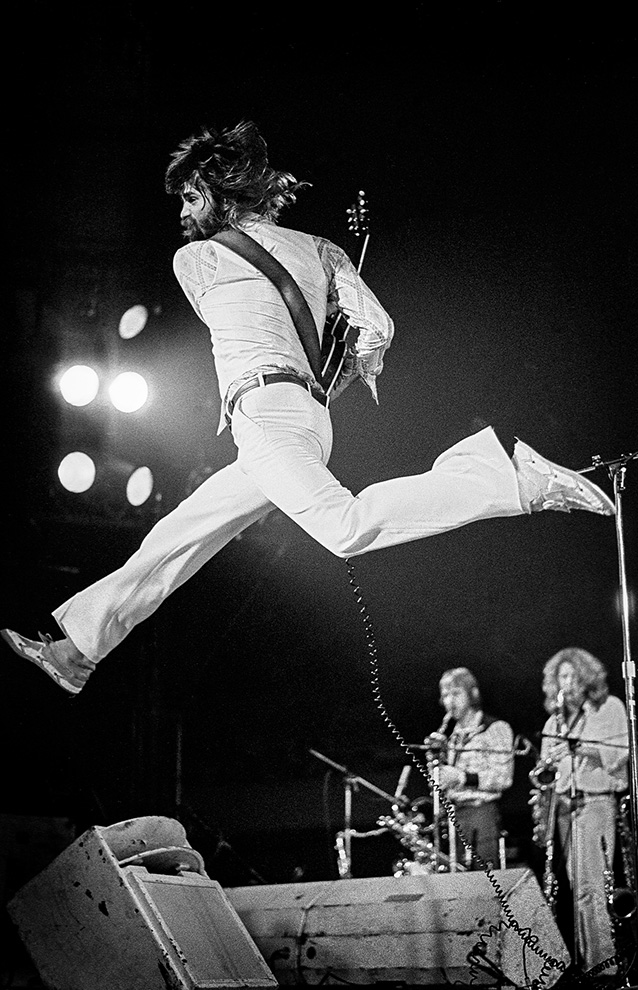 a person jumping mid air with a guitar, wearing a jumpsuit on stage