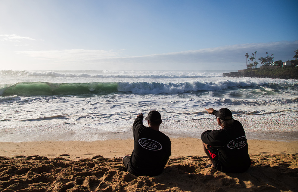 two people sitting on a beach shore watching the ocean waves